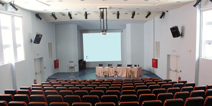 The main auditorium of the Agricultural University of Athens conference centre: tiered rows of orange and black seats facing a stage with a projection screen and a panel table.